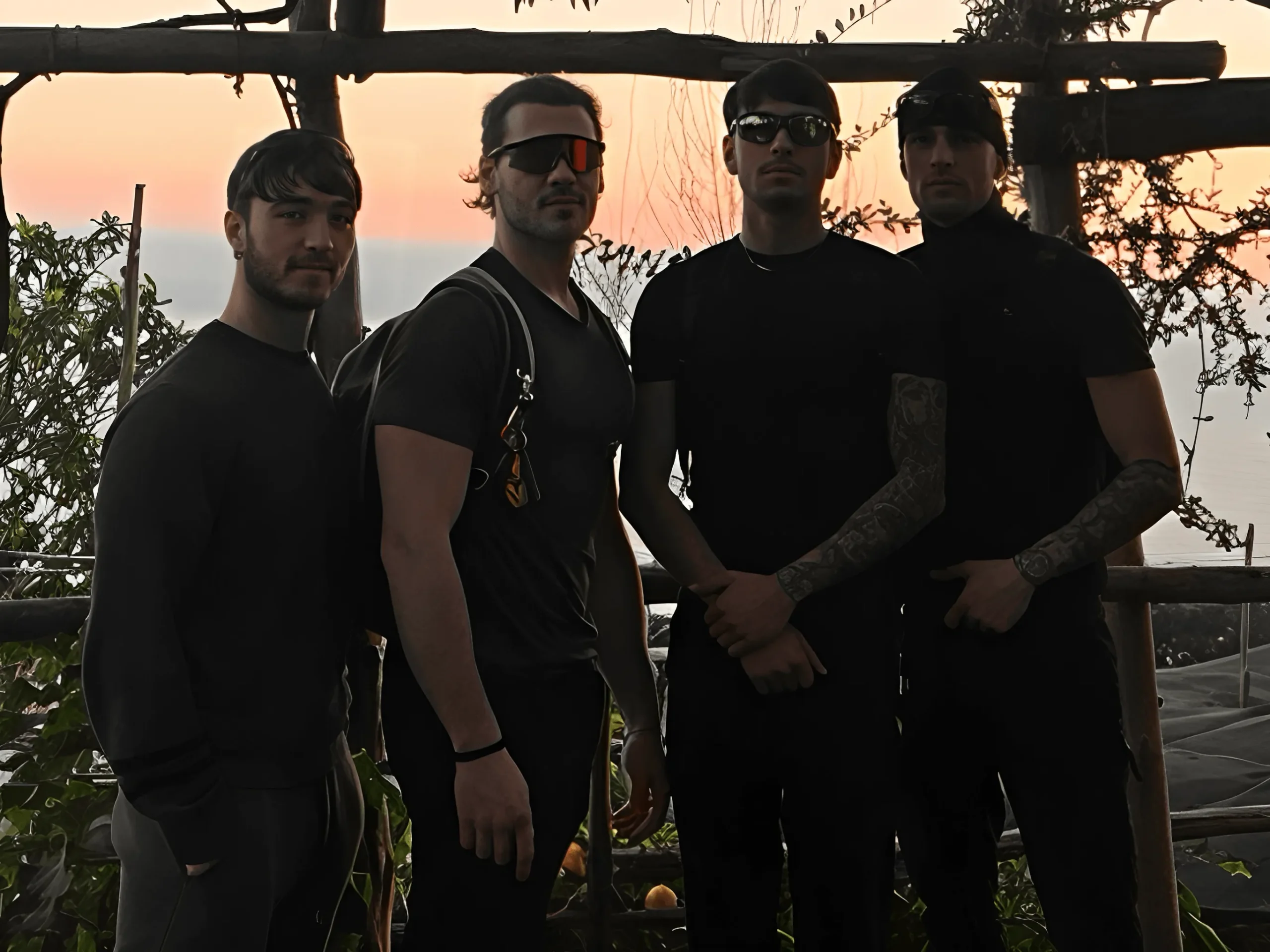 Four men stand outdoors at sunset, with trees and railings behind them. Three wear dark clothing and sunglasses; one wears a lighter shirt. The orange-hued sky and distant water set the scene for this Amalfi Outdoor Experience about us moment.