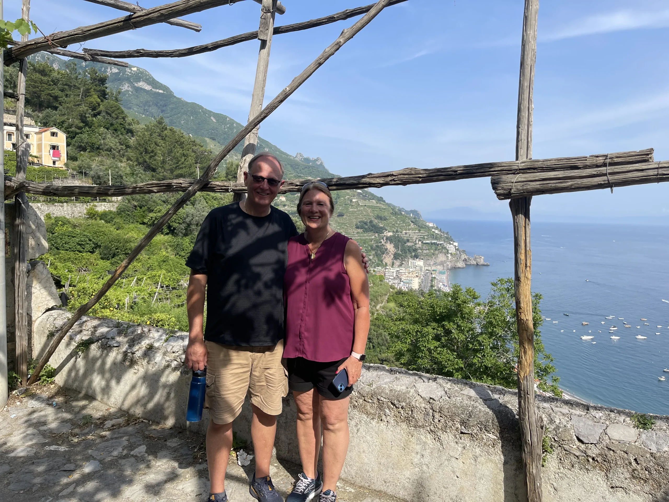 A smiling couple stands under a rustic wooden frame after hiking between Maiori and Minori, with lush green hills and a coastal village overlooking the sea in the background on a sunny day.