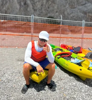Danilo, con un cappello bianco, occhiali da sole, giubbotto di salvataggio rosso e pantaloncini blu, è seduto sulla pagaia di un kayak giallo e punta la telecamera. Dietro di lui, sul terreno roccioso, sono appoggiati dei kayak colorati, con una recinzione e una parete rocciosa sullo sfondo. Danilo, con un cappello bianco, occhiali da sole, giubbotto di salvataggio rosso e pantaloncini blu, è seduto sulla pagaia di un kayak giallo e punta la telecamera. Dietro di lui, sul terreno roccioso, sono appoggiati dei kayak colorati, con una recinzione e una parete rocciosa sullo sfondo.