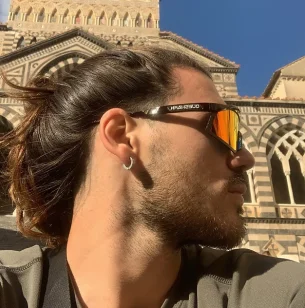 Federico, un uomo con i capelli lunghi, che indossa occhiali da sole catarifrangenti e un orecchino a cerchio, guarda di lato, davanti a un edificio in pietra ornato, a strisce, con archi e motivi geometrici, sotto un cielo azzurro. Federico, un uomo con i capelli lunghi, che indossa occhiali da sole catarifrangenti e un orecchino a cerchio, guarda di lato, davanti a un edificio in pietra ornato, a strisce, con archi e motivi geometrici, sotto un cielo azzurro.