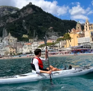 Un uomo in kayak pagaia sull'acqua limpida vicino alla vivace città costiera di Nicola, con edifici colorati e colline verdi sullo sfondo in una giornata di sole. Un uomo in kayak pagaia sull'acqua limpida vicino alla vivace città costiera di Nicola, con edifici colorati e colline verdi sullo sfondo in una giornata di sole.