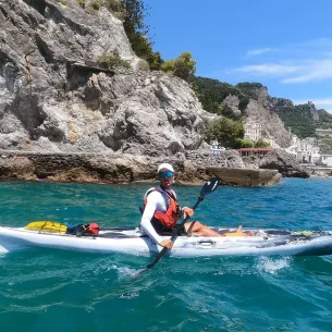 Salvatore, con occhiali da sole, cappello e giubbotto di salvataggio, sta facendo kayak su un'acqua azzurra e limpida, vicino a una costa rocciosa con scogliere scoscese e verde, sotto un cielo soleggiato. Salvatore, con occhiali da sole, cappello e giubbotto di salvataggio, sta facendo kayak su un'acqua azzurra e limpida, vicino a una costa rocciosa con scogliere scoscese e verde, sotto un cielo soleggiato.