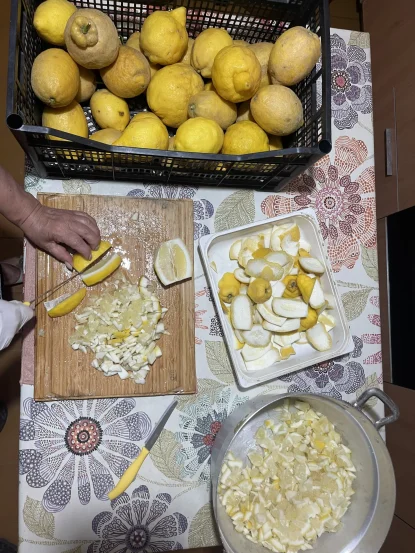 A person chops lemons on a wooden board near a basket of whole lemons, a tray with lemon peels, and a bowl of chopped lemon pieces—all set on a floral tablecloth reminiscent of picnics after the best hikes Amalfi Coast offers.