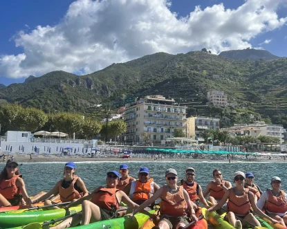 A group of people in orange life vests and hats sit in colorful kayaks on the water, smiling at the camera. Behind them are a beach, buildings near lemon path stairs, and green mountains under a partly cloudy sky with a sea view.