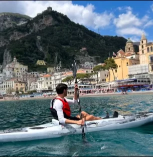 A person in a white kayak paddles near a coastal town with colorful buildings, a church—possibly dedicated to Saint Nicola—and lush green hills in the background under a partly cloudy sky.
