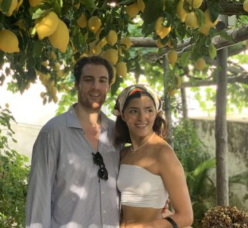A couple stands smiling under a pergola with hanging lemons and green leaves, evoking a sfusato amalfitano lemon tour. The woman wears a white tube top, light pants, and headscarf; the man sports a light blue shirt as sunlight filters through the foliage.
